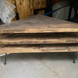 K&ouml;nnte beinhalten: Rustikales, dreieckiges Eckregal aus Holz mit drei offenen Ablagefl&auml;chen. Die Regalb&ouml;den sind aus dunkel gebeiztem Holz gefertigt und werden von d&uuml;nnen, schwarzen Metallbeinen getragen. Schlichtes Design.