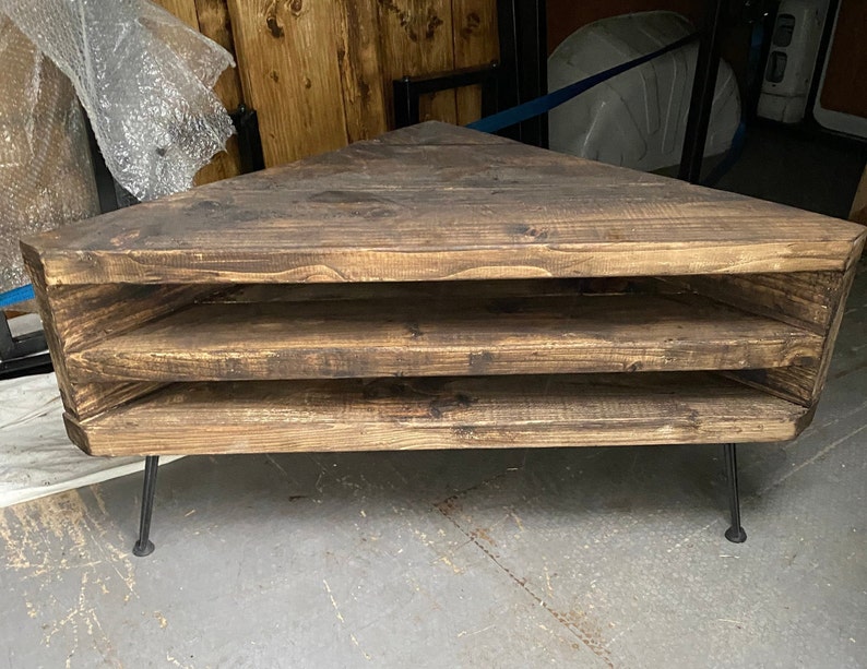K&ouml;nnte beinhalten: Rustikales, dreieckiges Eckregal aus Holz mit drei offenen Ablagefl&auml;chen. Die Regalb&ouml;den sind aus dunkel gebeiztem Holz gefertigt und werden von d&uuml;nnen, schwarzen Metallbeinen getragen. Schlichtes Design.