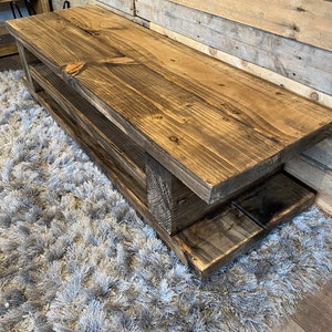 Op de afbeelding: Een rustieke houten bank met een donkere afwerking en twee onderste planken. De bank is gemaakt van dikke houten planken en staat op een grijze, harige vloerbedekking. De achtergrond is een houten wand.
