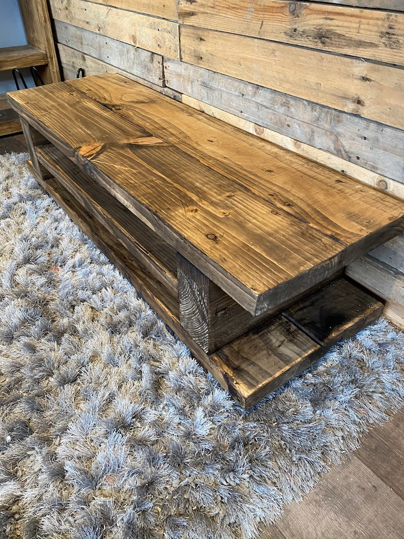 Op de afbeelding: Een rustieke houten bank met een donkere afwerking en twee onderste planken. De bank is gemaakt van dikke houten planken en staat op een grijze, harige vloerbedekking. De achtergrond is een houten wand.