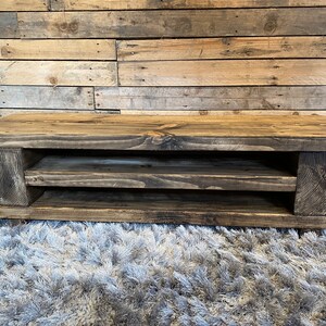 Op de afbeelding: Een rustieke, rechthoekige salontafel van donker gebeitst hout. De tafel heeft een onderste plank en wordt ondersteund door dikke, vierkante poten. De houtnerf is zichtbaar, wat het een natuurlijk, verouderd uiterlijk geeft. De tafel is ongeveer 122 cm lang.