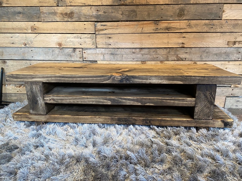 Op de afbeelding: Een rustieke, rechthoekige salontafel van donker gebeitst hout. De tafel heeft een onderste plank en wordt ondersteund door dikke, vierkante poten. De houtnerf is zichtbaar, wat het een natuurlijk, verouderd uiterlijk geeft. De tafel is ongeveer 122 cm lang.
