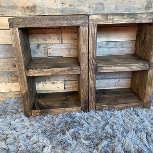 Op de afbeelding: Twee rustieke, donker gebeitste houten planken met elk twee niveaus. De planken zijn rechthoekig en lijken handgemaakt. De houtnerf is zichtbaar. De planken staan op een grijze, harige vloerkleed.