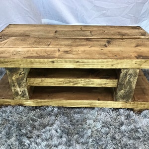 Op de afbeelding: Een rechthoekige salontafel van lichtbruin gerecycled hout. De tafel heeft een onderste plank en wordt ondersteund door dikke, stevige poten. Het hout heeft zichtbare nerven en knopen, wat het een natuurlijk, verouderd uiterlijk geeft. Ongeveer 120 cm lang.