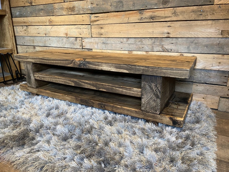 Op de afbeelding: Een rustieke, donker gebeitste houten tv-standaard met twee open planken. De standaard is gemaakt van dikke houten planken, die de natuurlijke nerf en knopen laten zien. Hij staat op een zacht, grijs tapijt, met een houten wand op de achtergrond.