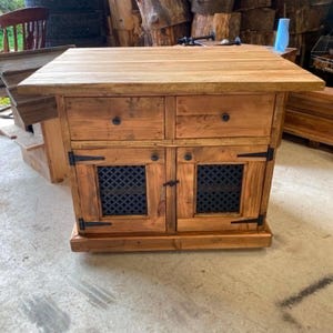 May include: A rectangular wooden kitchen island featuring a natural brown finish, two top drawers with small dark knobs, and two lower cabinet doors with black metal hinges and black decorative lattice inserts. The unit has a flat, planked top surface.