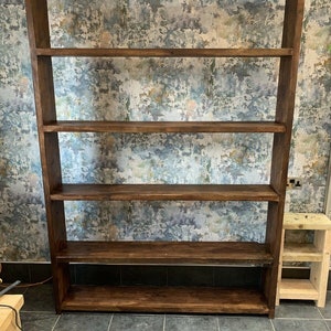 Op de afbeelding: Een hoge, donkerbruine houten boekenkast met vijf planken. De planken zijn gelijkmatig verdeeld en het hout heeft een natuurlijke nerf. De boekenkast is vrijstaand. Een kleine, lichtgekleurde houten bijzettafel staat naast de boekenkast. De boekenkast is ongeveer 180 cm hoog.