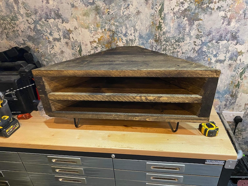 K&ouml;nnte beinhalten: Rustikales, dreieckiges Eckregal aus dunkel gebeiztem Holz. Es hat zwei Innenb&ouml;den und wird von schwarzen Haarnadelbeinen getragen. Das Regal ist so konzipiert, dass es perfekt in eine Ecke passt und Platz spart.