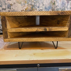 K&ouml;nnte beinhalten: Rustikaler Eck-TV-Schrank aus Holz mit zwei Regalen und Haarnadelbeinen. Das helle Holz zeigt Maserung und Astl&ouml;cher. Das M&ouml;belst&uuml;ck ist f&uuml;r Ecken konzipiert und bietet Stauraum. Ma&szlig;e in Zentimetern.