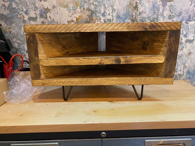 K&ouml;nnte beinhalten: Rustikaler Eck-TV-Schrank aus Holz mit zwei Regalen und Haarnadelbeinen. Das helle Holz zeigt Maserung und Astl&ouml;cher. Das M&ouml;belst&uuml;ck ist f&uuml;r Ecken konzipiert und bietet Stauraum. Ma&szlig;e in Zentimetern.