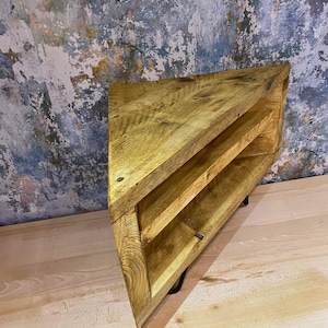 K&ouml;nnte beinhalten: Eckregal aus Holz mit nat&uuml;rlicher Oberfl&auml;che. Das Regal hat eine dreieckige Form mit zwei Innenb&ouml;den. Die Holzmaserung ist sichtbar und das Regal wird von schwarzen Metallbeinen getragen. Der Hintergrund zeigt eine abgenutzte Wand.