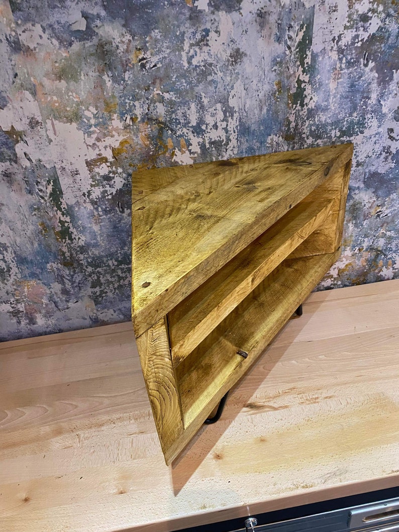 K&ouml;nnte beinhalten: Eckregal aus Holz mit nat&uuml;rlicher Oberfl&auml;che. Das Regal hat eine dreieckige Form mit zwei Innenb&ouml;den. Die Holzmaserung ist sichtbar und das Regal wird von schwarzen Metallbeinen getragen. Der Hintergrund zeigt eine abgenutzte Wand.