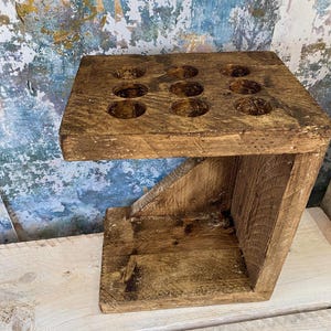 May include: A wooden shot glass holder with nine circular recesses. The holder is made from reclaimed wood, with a natural, weathered appearance. It features a lower shelf and an angled support, showcasing a rustic design.