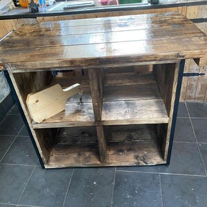 May include: A rectangular kitchen island crafted from dark stained wood with a glossy finish. The unit features a flat top surface and four open storage compartments arranged in a two-by-two grid. A light-coloured wooden cutting board rests inside.