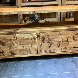 Op de afbeelding: Een rustieke tv-standaard van natuurlijk hout. Het heeft twee lades met ronde houten knoppen en zwarte metalen scharnieren. Boven de lades bevindt zich een open plank. Het meubel is gemaakt van massief hout en is ongeveer 150 cm breed.