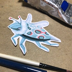 May include: A paper cutout of a blue and red spotted bug with white accents. The bug has six legs and a pair of antennae.