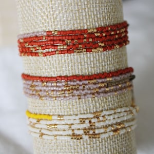 Peut inclure: Un ensemble de bracelets en perles colorées sur un présentoir en tissu beige. Les bracelets sont faits de petites perles rondes dans des tons de rouge, blanc et or.