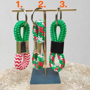 May include: Three green, white and pink rope keychains with gold and black accents hanging on a gold stand. The keychains are numbered 1, 2 and 3.