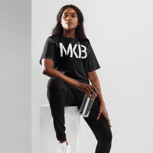 May include: A person wearing a black t-shirt with the word "MK3" printed in white on the front. The person is sitting on a white surface and holding a clear water bottle.