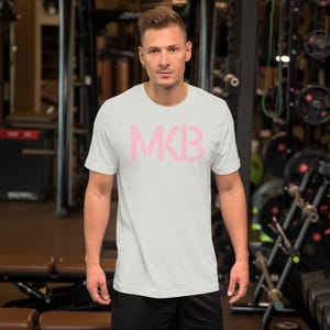 May include: White t-shirt with a pink "MKB" logo printed on the front. The shirt is being worn by a person in a gym setting.