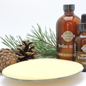 May include: An amber glass bottle and a smaller dropper bottle with the text "Pollen de" and "Pollen de pin". A pine cone, pine needles, and a light yellow dish are also in the image. The bottles have a brown cap and a label with a geometric design.