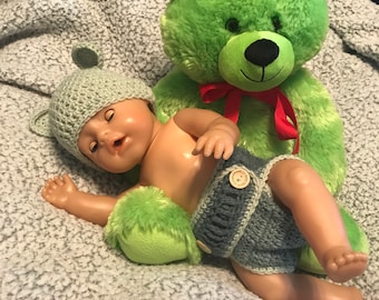 May include: A green plush teddy bear with a red bow sits next to a baby doll wearing a gray knitted hat and a blue and white knitted diaper. The baby doll is lying on a white fluffy blanket.