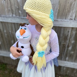 Könnte beinhalten: Ein junges Mädchen, verkleidet als Elsa aus Frozen, trägt eine gelbe Häkelmütze mit einem geflochtenen gelben Garnzopf und blauen Schneeflockenverzierungen. Sie hält eine Plüsch-Olaf-Puppe.