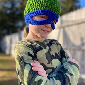 May include: A child wearing a green and blue crocheted hat with a mask. The hat has a blue eye mask with a green brim. The child is wearing a camouflage shirt and has their arms crossed.