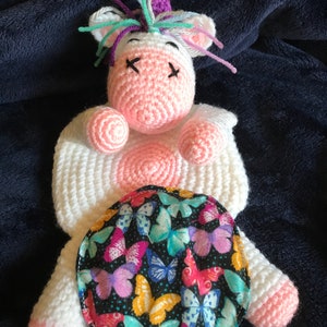 May include: A white crocheted unicorn stuffed animal with pink accents and a rainbow mane. The unicorn is lying down with its eyes closed and has a round, black fabric patch with a butterfly pattern on its back.