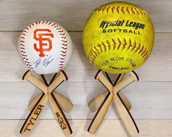 May include: Two baseballs on wooden baseball bat stands. The baseball on the left is white with an orange San Francisco Giants logo and a signature. The baseball on the right is yellow with the text "Official League Softball" and "12.2IN 44 COR 375CM YWC S12".