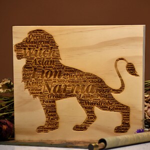 Pode incluir: Placa de madeira clara com um desenho de leão esculpido, preenchido com palavras como "Nárnia" e "Aslan". O design é realçado por flores secas e um pergaminho. O fundo é castanho.