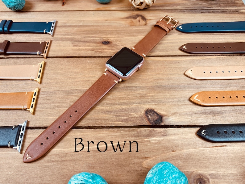 May include: A brown leather Apple Watch band with a rose gold Apple Watch on a wooden surface. The band is shown with other leather bands in different colors.