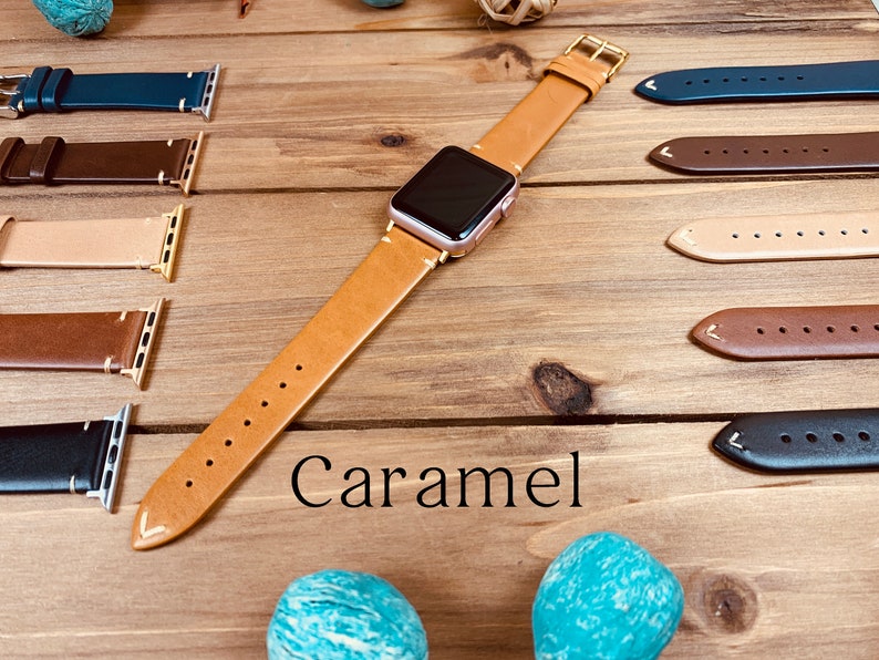 May include: Caramel leather Apple Watch band with a gold buckle. The band is shown on a wooden surface with other leather bands in different colors.