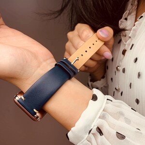May include: A navy blue leather watch band with a tan leather lining and stitching. The band is attached to a rose gold smartwatch.