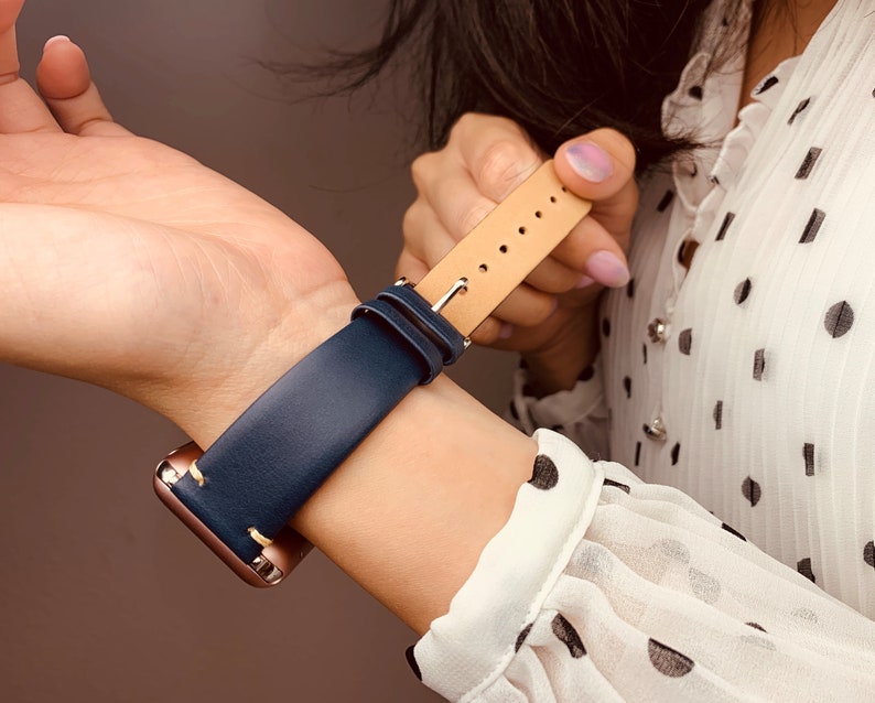 May include: A navy blue leather watch band with a tan leather lining and stitching. The band is attached to a rose gold smartwatch.