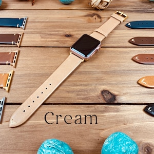May include: A cream-colored leather watch band for an Apple Watch. The band is shown laid out on a wooden surface with other watch bands in various colors. The text "Cream" is printed below the band.