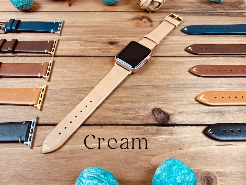 May include: A cream-colored leather watch band for an Apple Watch. The band is shown laid out on a wooden surface with other watch bands in various colors. The text "Cream" is printed below the band.