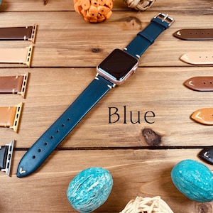 May include: A blue leather watch band with a rose gold watch face. The band is laid out on a wooden surface with other watch bands in different colors.