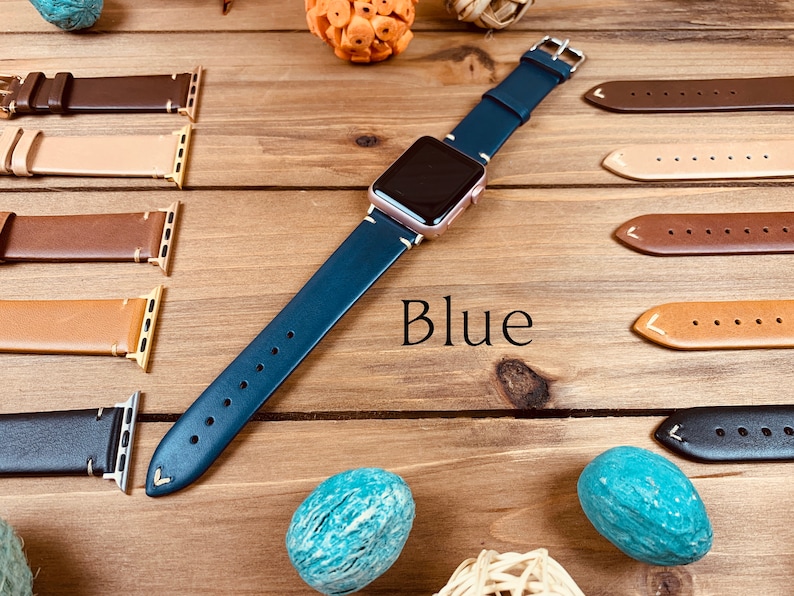 May include: A blue leather watch band with a rose gold watch face. The band is laid out on a wooden surface with other watch bands in different colors.