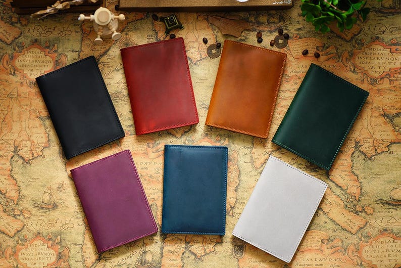 May include: Seven leather passport holders in different colours, including black, red, brown, green, blue, purple and white, are arranged on a map background.