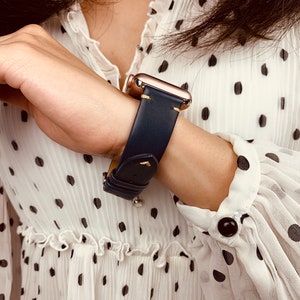 May include: A navy blue leather watch band with gold hardware and stitching. The band is worn on a wrist with a white polka dot shirt.