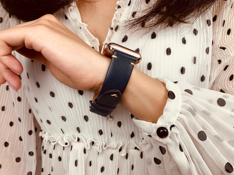 May include: A navy blue leather watch band with gold hardware and stitching. The band is worn on a wrist with a white polka dot shirt.
