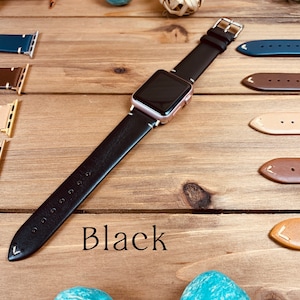 May include: A black leather Apple Watch band with a rose gold watch face. The band is shown on a wooden surface with other leather bands in different colors.