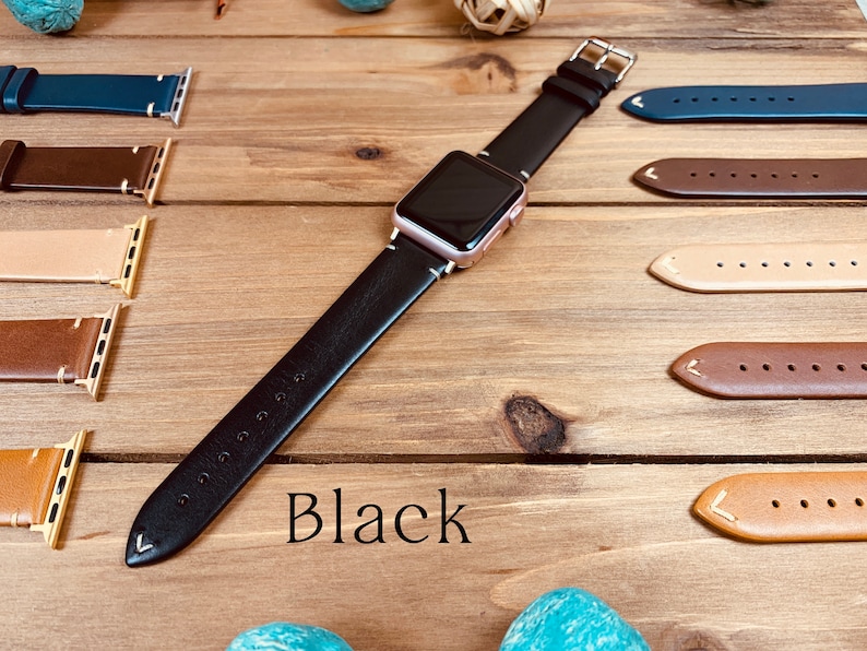 May include: A black leather Apple Watch band with a rose gold watch face. The band is shown on a wooden surface with other leather bands in different colors.