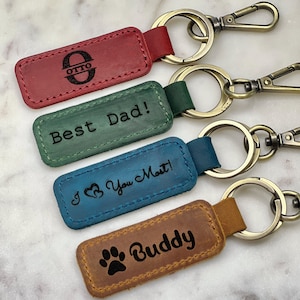May include: Four leather keychains with different engravings. The keychains are red, green, blue, and brown. The engravings include "Otto", "Best Dad!", "I Love You Most!" and "Buddy".