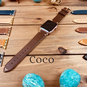 May include: A brown leather watch band with a rose gold watch face. The band has a stitched edge and a pointed end. The watch band is displayed on a wooden surface with other watch bands in different colors.