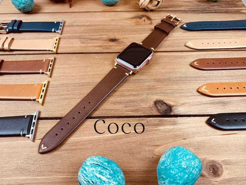 May include: A brown leather watch band with a rose gold watch face. The band has a stitched edge and a pointed end. The watch band is displayed on a wooden surface with other watch bands in different colors.