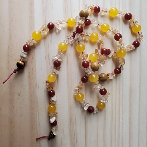 Op de afbeelding: Een lange ketting gemaakt van gele, oranje en rode edelsteen kralen. De ketting is gemaakt van kralen in verschillende vormen en maten, waaronder ronde, ovale en onregelmatige vormen. De ketting is geregen op een rood koord.
