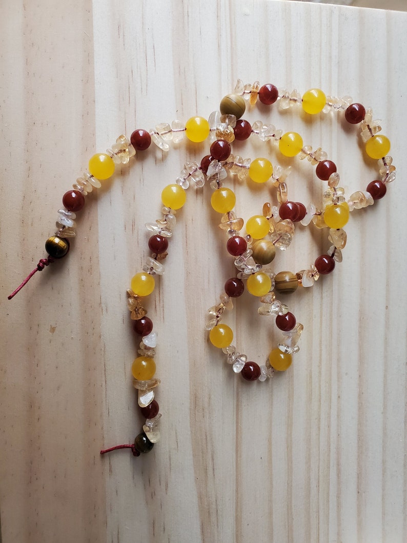 Op de afbeelding: Een lange ketting gemaakt van gele, oranje en rode edelsteen kralen. De ketting is gemaakt van kralen in verschillende vormen en maten, waaronder ronde, ovale en onregelmatige vormen. De ketting is geregen op een rood koord.