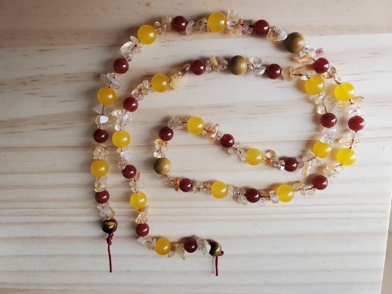 Op de afbeelding: Een lange ketting gemaakt van gele, oranje en rode edelsteen kralen. De ketting is gemaakt van kralen in verschillende maten en vormen, waaronder enkele kleine, onregelmatige stukjes. De ketting heeft twee tijgeroog kralen aan de uiteinden.